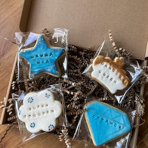 May include: Four decorated sugar cookies in a box. The cookies are shaped like a star, a crown, a flower, and a diamond. The star is blue with gold glitter and says "Mamma Mia". The crown is white with gold glitter and says "Dancing Queen". The flower is white with blue and yellow flowers and says "Super Trouper". The diamond is blue with gold glitter and says "Dynamos".