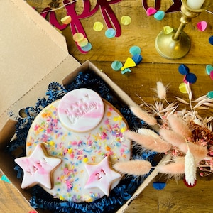 Pu&ograve; includere: Una scatola regalo di compleanno con una torta rotonda decorata con codette colorate e una stella rosa e bianca con il numero 21. La scatola contiene anche una stella rosa e bianca con le parole "YAY". La scatola &egrave; circondata da coriandoli colorati e uno striscione rosso che dice "Happy".