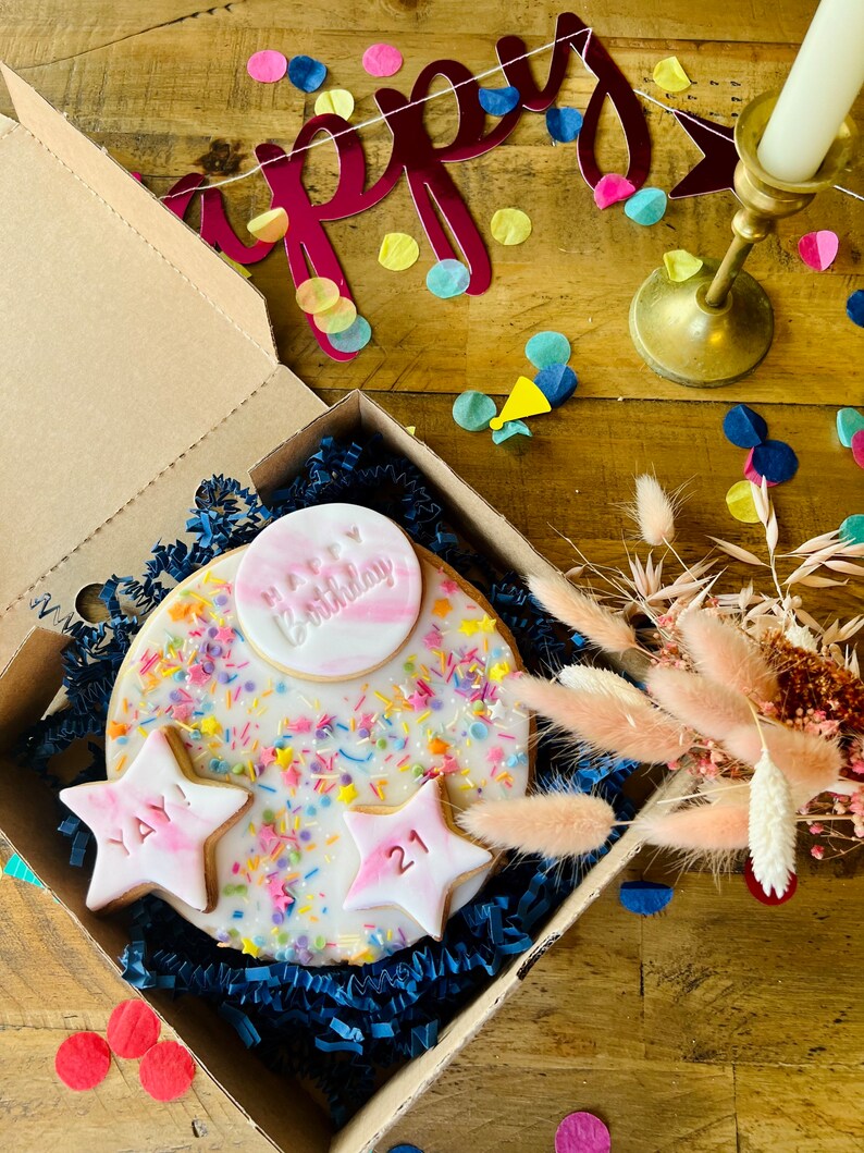 Pu&ograve; includere: Una scatola regalo di compleanno con una torta rotonda decorata con codette colorate e una stella rosa e bianca con il numero 21. La scatola contiene anche una stella rosa e bianca con le parole "YAY". La scatola &egrave; circondata da coriandoli colorati e uno striscione rosso che dice "Happy".