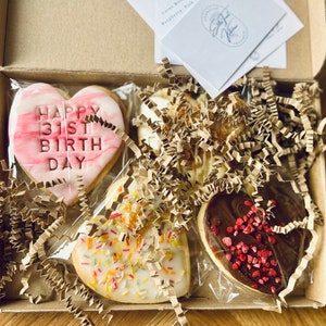 Puede incluir: Una caja de regalo llena de cuatro galletas con forma de coraz&oacute;n. Una galleta es rosa con vetas blancas y dice "Happy 31st Birthday". Otra galleta es blanca con chispas de colores. La tercera galleta es blanca con una cobertura de chocolate marr&oacute;n y chispas rojas. La cuarta galleta es marr&oacute;n con chispas rojas.