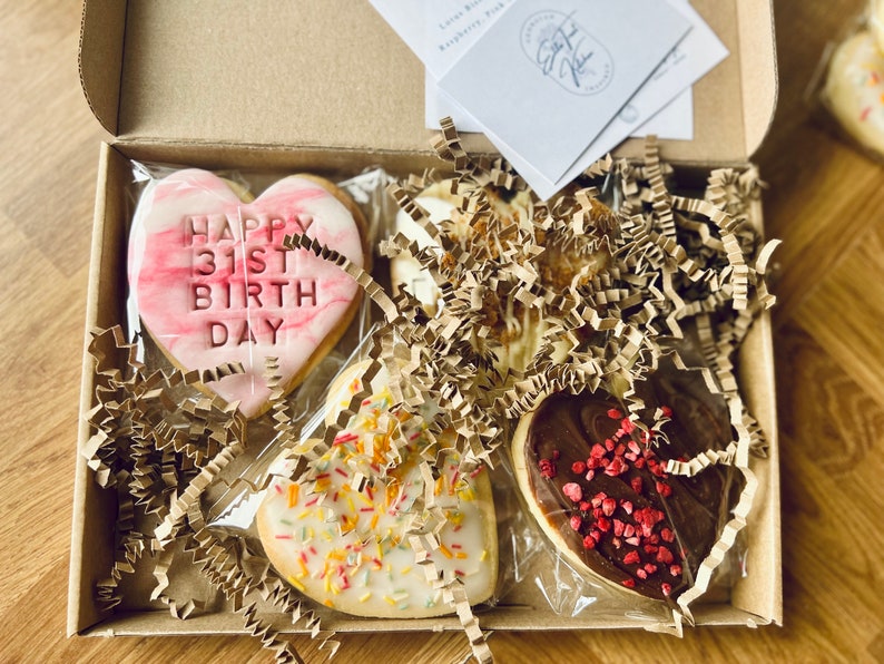 Puede incluir: Una caja de regalo llena de cuatro galletas con forma de coraz&oacute;n. Una galleta es rosa con vetas blancas y dice "Happy 31st Birthday". Otra galleta es blanca con chispas de colores. La tercera galleta es blanca con una cobertura de chocolate marr&oacute;n y chispas rojas. La cuarta galleta es marr&oacute;n con chispas rojas.
