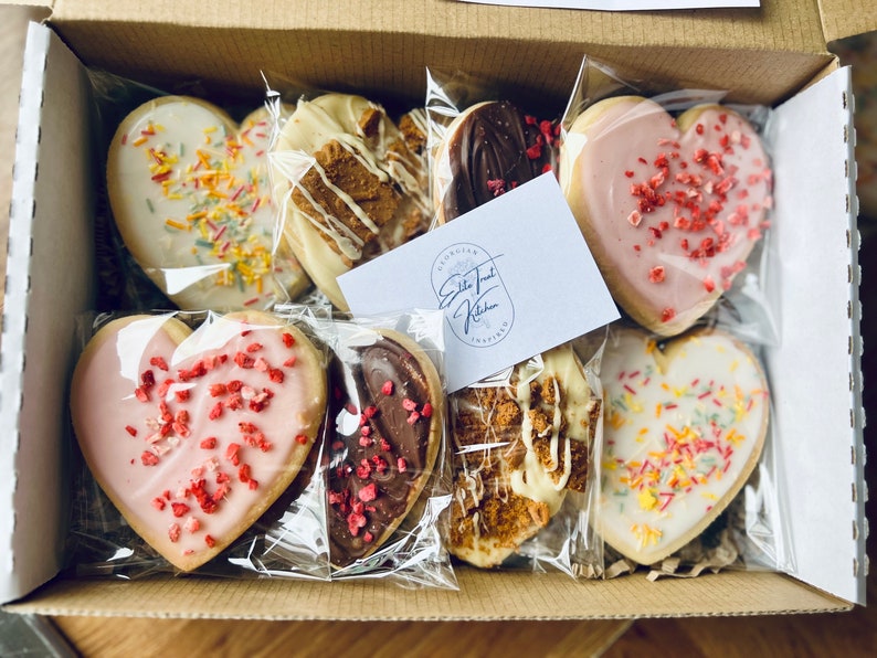 Puede incluir: Una caja de galletas surtidas en forma de coraz&oacute;n, que incluyen chocolate, chocolate blanco y chispas. Las galletas est&aacute;n envueltas individualmente en bolsas de pl&aacute;stico transparentes. La caja est&aacute; etiquetada como "Georgian Sweet Treat Kitchen Inspired".