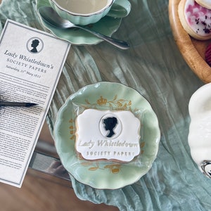 K&ouml;nnte beinhalten: Nahaufnahme eines verzierten Zuckerkekses mit dem Text "Lady Whistledown's Society Papers" in goldenen Lettern. Der Keks liegt auf einem gr&uuml;n-goldenen Unterteller mit Blumenmuster.
