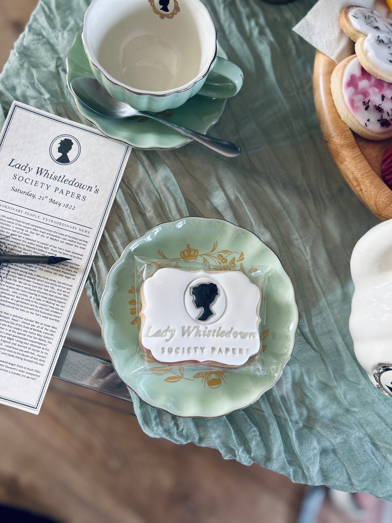 K&ouml;nnte beinhalten: Nahaufnahme eines verzierten Zuckerkekses mit dem Text "Lady Whistledown's Society Papers" in goldenen Lettern. Der Keks liegt auf einem gr&uuml;n-goldenen Unterteller mit Blumenmuster.