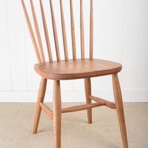 May include: A solid wood chair with a light brown finish. The chair has a curved back with vertical slats and a simple, rectangular seat. The chair has four legs and a stretcher connecting the front legs.