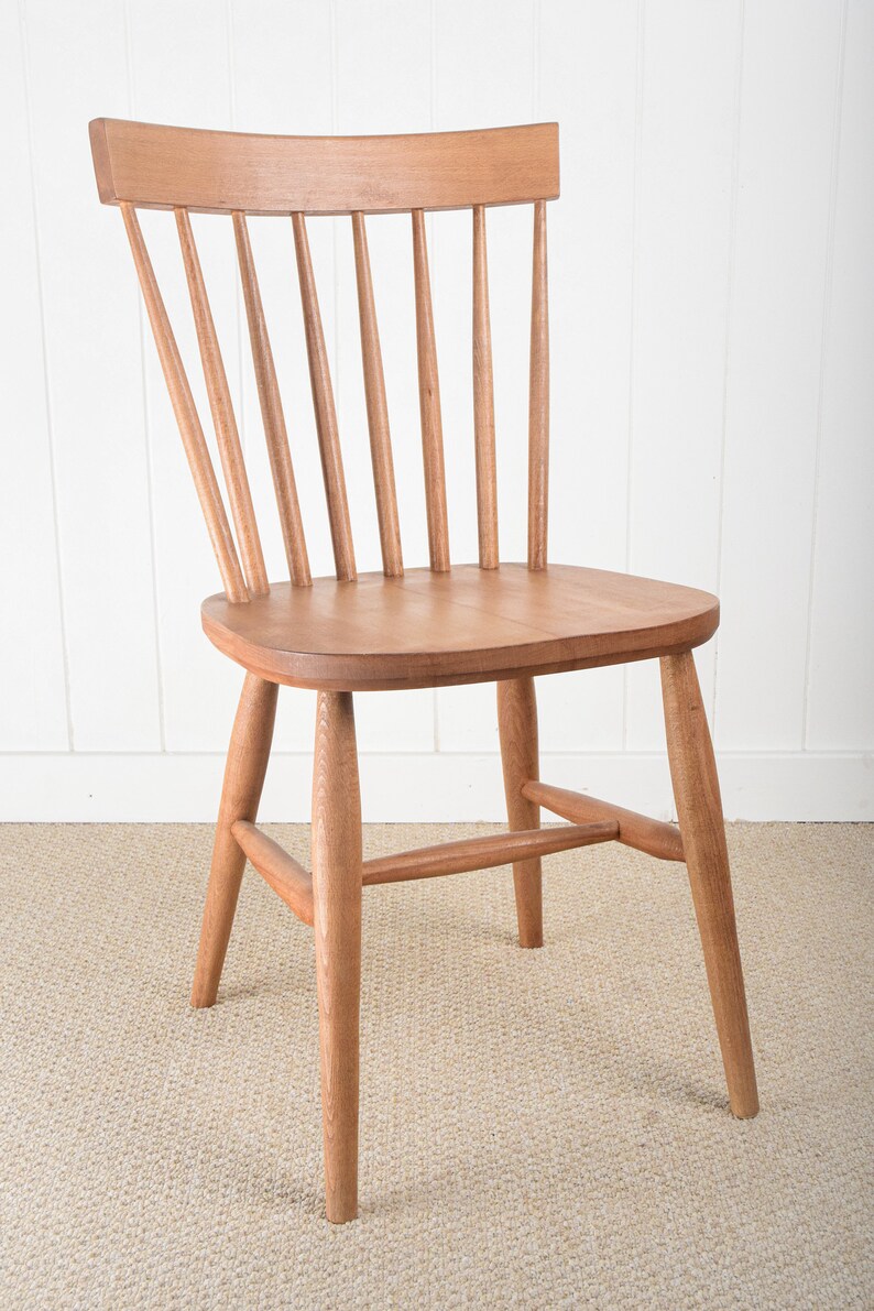 May include: A solid wood chair with a light brown finish. The chair has a curved back with vertical slats and a simple, rectangular seat. The chair has four legs and a stretcher connecting the front legs.