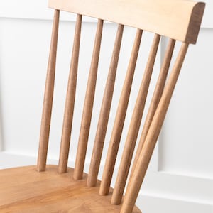 May include: Close-up of the back of a wooden chair with a slatted back. The chair is made of light brown wood and has a simple, classic design.
