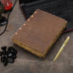 May include: A brown leather journal with aged, yellowed pages. The journal is open, revealing the texture of the paper. A gold pen with a textured design rests beside the journal. Black polished stones are scattered nearby.