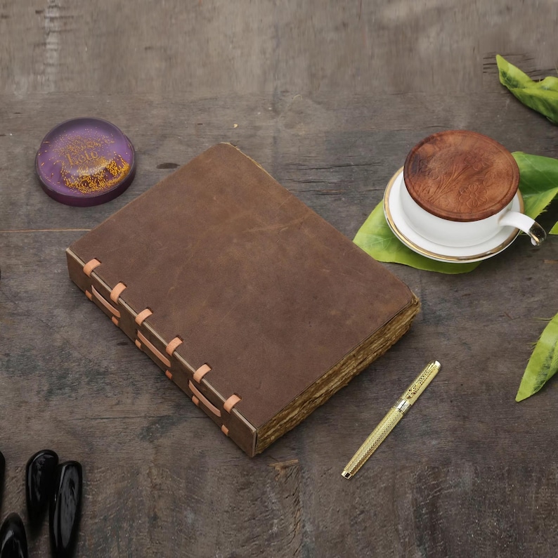 May include: A brown leather journal with stitched binding sits on a wooden surface. A gold pen, a cup of coffee, and a purple paperweight are also present. The journal has aged, yellowed pages, suggesting it is vintage.