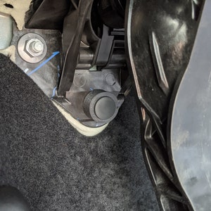 Puede incluir: Primer plano de una tapa de plástico negro unida a un panel interior de un coche. La tapa está situada cerca de una alfombra negra y un tornillo de metal.