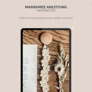 May include: A wooden-topped pacifier clip with a cream-colored macrame design, displayed on a wooden surface. The image includes text in German: "MAKRAMEE ANLEITUNG HERZKNOTEN" and "Süße Schnullerkette zum selber Knoten!" Also, a sprig of dried flowers.