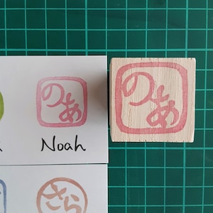 May include: A wooden block stamp with a pink ink impression of the Japanese character for "Noah". The stamp is on a green grid background and a white card with the name "Noah" written in black ink.