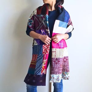 以下が含まれることがあります： カラフルな布の切れ端で作られたパッチワークのストール。ストールはピンク、パープル、ブルー、レッド、ホワイトの布のミックスです。ストールは、黒いシャツとジーンズを着た人の首に巻かれています。