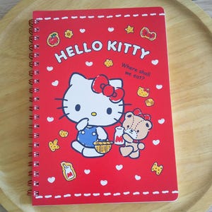 May include: A red spiral-bound notebook featuring Hello Kitty and a teddy bear on the cover. The text reads HELLO KITTY and Where shall we eat? The design includes white hearts, small food icons, and a dashed border along the top and bottom edges.