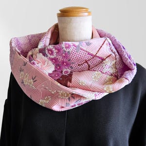 May include: A pleated infinity scarf featuring a light pink base with a repeating pattern of white and dark pink floral blossoms, geometric grid sections, and deep purple accents. The fabric has a textured, crinkled surface throughout the loop.