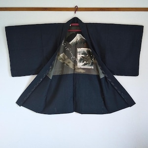 以下が含まれることがあります： 濃いネイビーブルーの着物が開いて吊るされており、山の風景が描かれた模様の裏地が見えます。着物は赤いハンガーに吊るされ、白い壁を背景にしています。袖は幅広く、生地は柔らかく見えます。