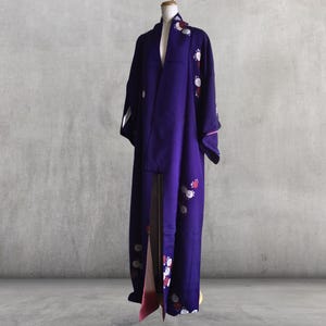 May include: A long, flowing purple kimono with floral embroidery in white, red, and pink. The kimono is displayed on a mannequin against a gray background. The sleeves and hem are adorned with the floral pattern.