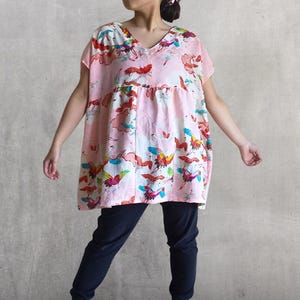 May include: A light pink tunic top with a V-neck and short sleeves. The top features a colorful bird and cloud pattern. The model is wearing dark blue pants.