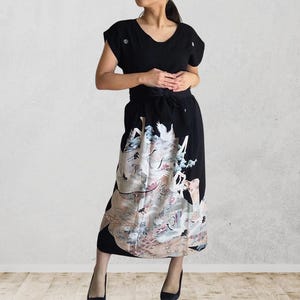 May include: Black dress with a V-neck and short sleeves. The skirt features a detailed, colorful illustration with birds and floral elements. The dress has a tie-waist design and is paired with black heels.