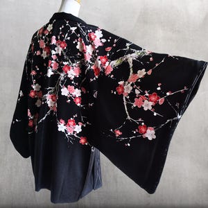 以下が含まれることがあります： 赤、ピンク、白の梅の花が咲く枝のプリントが施された黒い羽織。この羽織は、幅の広い袖としっかりとした襟が特徴で、マネキンに着せられた状態で撮影されています。全体的に落ち着いた色合いで、花柄が鮮やかに描かれています。