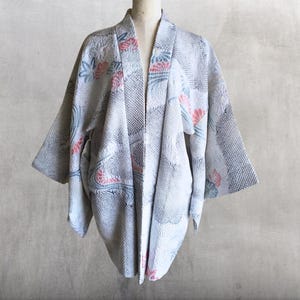 以下が含まれることがあります： ピンクとブルーの花柄が施された、ライトグレーの着物風ジャケット。 ジャケットは袖が広く、テクスチャード加工された生地です。 この衣服は、ニュートラルな背景のマネキンに展示されています。