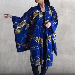 以下が含まれることがあります： 鮮やかな黄色、白、緑の花柄と風景模様が施された、長く流れるような青色の着物。 着物はゆったりとしたフィット感で、快適でスタイリッシュな外観を提供します。 この着物は軽量の生地で作られています。