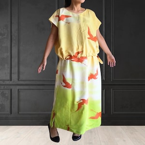 May include: A yellow dress with a crane print. The dress has a round neckline, short sleeves, and a gathered waist. The skirt has a green and white gradient with orange cranes. The dress is knee-length.
