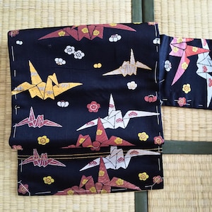 May include: A folded black kimono with a repeating pattern of origami cranes in yellow, red, and white, along with small floral and geometric designs. The kimono is displayed on a woven mat.