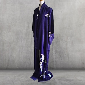 May include: A dark purple kimono with floral embroidery. The kimono is displayed on a mannequin against a gray background. The floral embroidery is white and light pink. The kimono is long and flowing.