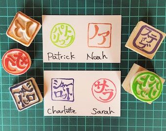 May include: A set of five colorful rubber stamps with Japanese characters and names written below each stamp. The names are Patrick, Noah, Charlotte, and Sarah. The stamps are on a green grid background.