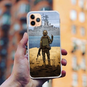 Puede incluir: Una funda para teléfono con un gráfico de un soldado de pie en una playa con un barco en el fondo. El soldado sostiene un rifle y mira hacia el mar. La funda es blanca con una parte posterior transparente.