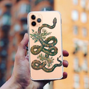Puede incluir: Una funda de iPhone dorada con un diseño de serpiente verde. La serpiente está enroscada alrededor de un grupo de hojas y flores verdes. La funda tiene una parte posterior transparente y una protuberancia de cámara negra.