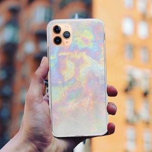 以下が含まれることがあります： 虹色の光沢のあるパターンが施された白いiPhoneケース。ケースにはカメラレンズの切り抜きと透明な背面があります。