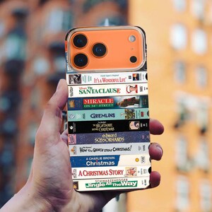 May include: An orange phone case displaying a stack of VHS tapes with festive film titles including "It's a Wonderful Life," "Gremlins," and "A Christmas Story." The case has a clear border and an orange camera module.