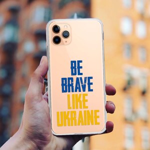 Könnte beinhalten: Eine transparente Handyhülle mit goldenem Hintergrund und dem Text "BE BRAVE LIKE UKRAINE" in blauen und gelben Buchstaben.