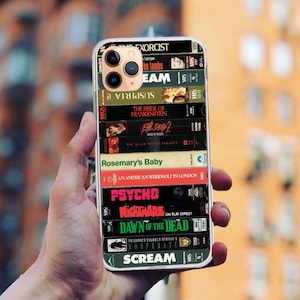 May include: A black iPhone case featuring a collage of horror movie VHS tapes. The tapes include titles such as "The Exorcist", "Silence of the Lambs", "Suspiria", "The Bride of Frankenstein", "Evil Dead 2", "Rosemary's Baby", "An American Werewolf in London", "Psycho", "Nightmare on Elm Street", "Dawn of the Dead", "Scream".