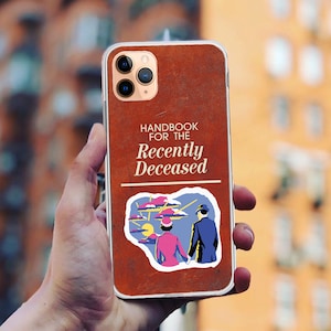 Peut inclure: Une coque de téléphone iPhone en cuir marron avec un autocollant qui dit "Handbook for the Recently Deceased". L'autocollant représente un couple dans un décor fantaisiste.