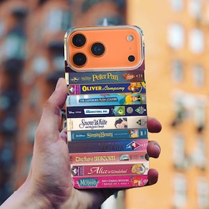 May include: A clear phone case with an orange camera module, decorated with a stack of colourful VHS tapes. Titles include "Peter Pan," "Oliver & Company," "Snow White," and "Alice in Wonderland." The case is held in a hand.