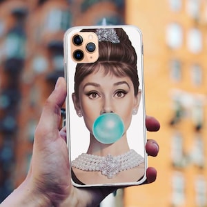 May include: A clear phone case with a white background and a colorful illustration of a woman with a pearl necklace blowing a bubble. The woman has dark hair styled in a bun and is wearing a pearl necklace.
