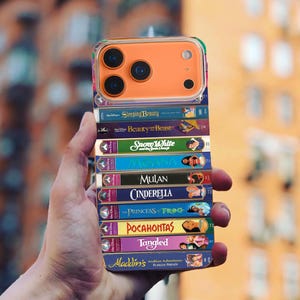 Puede incluir: Una funda transparente para teléfono con un módulo de cámara naranja, que presenta una pila de cintas VHS coloridas con títulos de películas de Disney como "Sleeping Beauty", "Beauty and the Beast" y "Tangled". La funda se sostiene contra un fondo urbano borroso.