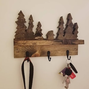 Può includere: Portachiavi in legno con una silhouette di una scena di foresta. Il portachiavi ha tre ganci ed è tinto di un colore marrone scuro.