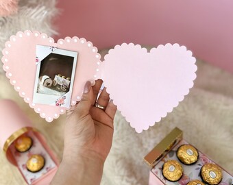 Può includere: Una scatola rosa a forma di cuore con un bordo a festoni, aperta per rivelare una scatola più piccola a forma di cuore con una foto all'interno. La scatola è decorata con pois bianchi e accenti floreali. La scatola più piccola è anche rosa e ha un bordo a festoni.