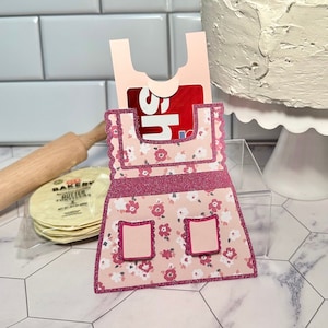 May include: A pink apron-shaped gift card holder with a floral pattern and glitter accents. The holder has two small square pockets and a scalloped edge. A gift card is partially visible in the top pocket. A rolling pin and tortillas are in the background.