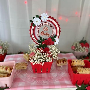May include: A table setting with a red berry basket filled with flowers and pastries. A circular sign with the text "Oh Baby" is placed above the basket. The arrangement sits on a clear plate over a pink gingham tablecloth.