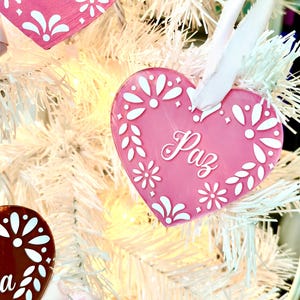 May include: Pink heart-shaped ornament with white floral designs and the word "Paz" written in white. The ornament is hanging on a white artificial Christmas tree.