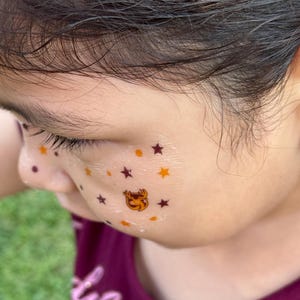 May include: Close-up of a child's face with temporary tattoos. The tattoos include small orange and brown stars, and a crab design. The child is wearing a maroon shirt.
