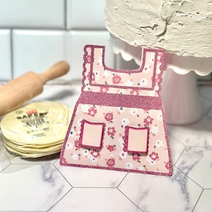 May include: A pink paper apron with a floral pattern and glitter detailing. The apron features two small pockets and a scalloped edge. A rolling pin and a stack of tortillas are in the background, suggesting a baking theme.