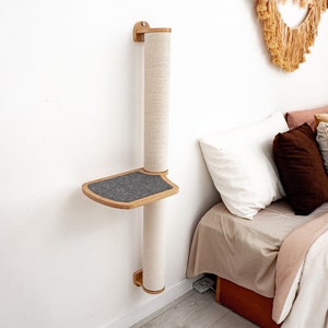 May include: A wall-mounted cat scratching post with a platform and sisal rope. The post is made of natural wood and has a grey platform. The post is mounted on a white wall next to a bed with pillows.
