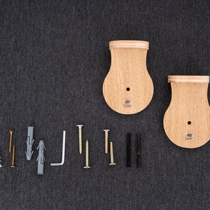 以下が含まれることがあります： 明るい木製の仕上げと丸い形をした木製の壁掛けが2つ。マウントの中央には小さな穴があり、下部に「Htt Haken」というテキストが印刷されています。画像には、さまざまなネジ、アンカー、金属製のL字型ブラケットも表示されています。
