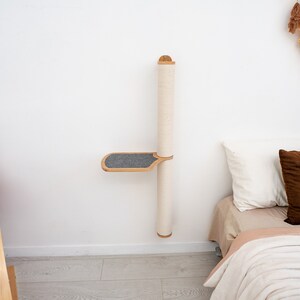 Pode incluir: Um arranhador de parede para gatos com uma plataforma. O poste &eacute; coberto com corda de sisal e a plataforma &eacute; coberta com tecido cinza. O poste &eacute; montado em uma parede branca e h&aacute; uma cama com almofadas no fundo.